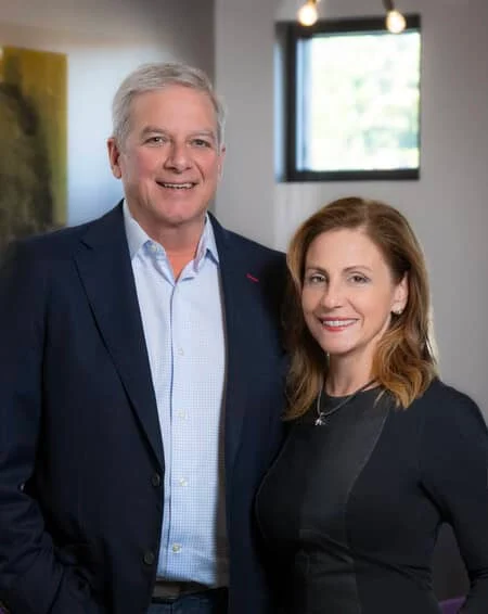 A man in a dark blazer and a woman in a fitted black dress stand together, smiling, in a well-lit indoor setting.