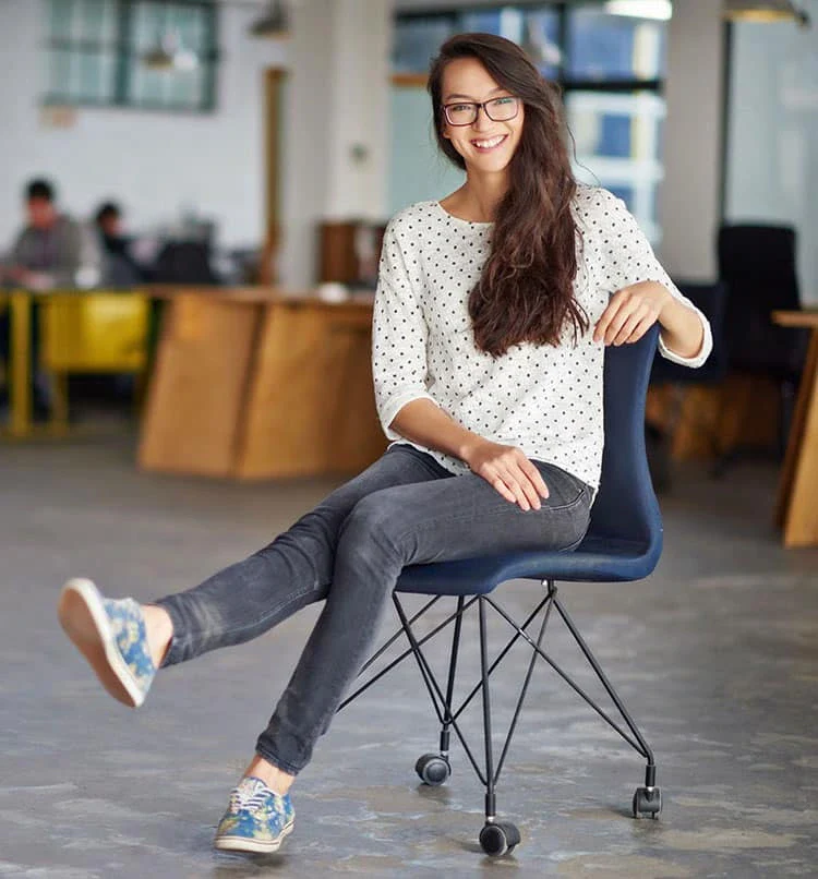 A woman sits casually on a modern chair in a bright, open workspace, wearing a dotted sweater and jeans, with colorful shoes.
