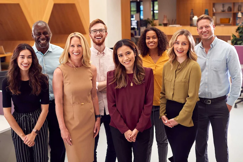 A diverse group of professionals stands together in a modern office setting, wearing a mix of casual and business attire.
