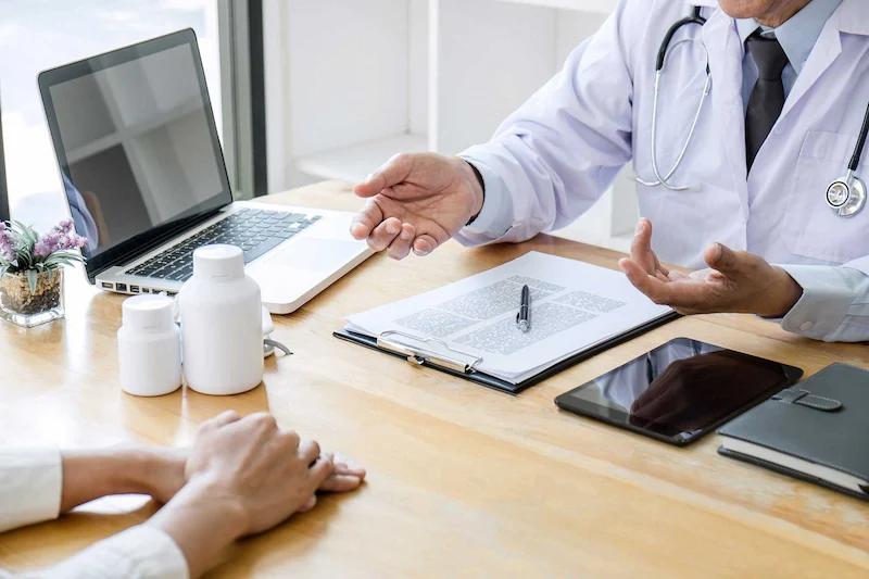 A doctor in a white coat discusses treatment options with a patient at a desk, with medication bottles and a laptop in view.