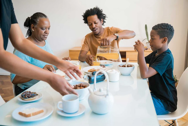 A family enjoys breakfast at a table, serving cereal, toast, and pouring juice, with a lively and inviting atmosphere.