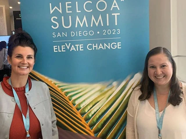 Two individuals stand in front of a blue backdrop promoting the WELCOA Summit 2023 in San Diego, emphasizing the theme - Elevate Change.