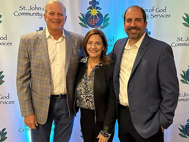 Three individuals pose for a photo in front of a backdrop featuring the St. John of God Community Services logo and decorative elements.