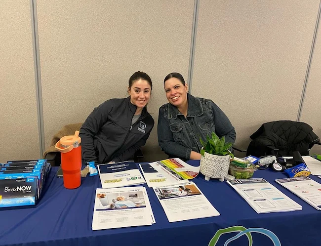 Two individuals sit closely behind a table filled with informational brochures, a plant, and various promotional materials.