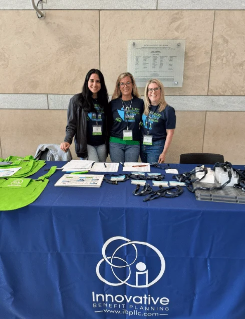 Three women stand behind a table displaying informational materials and green tote bags, representing Innovative Benefit Planning.