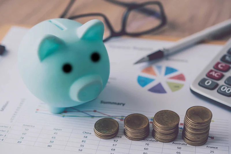 A blue piggy bank sits on financial documents beside stacked coins, a calculator, and a pen, suggesting budgeting and saving.