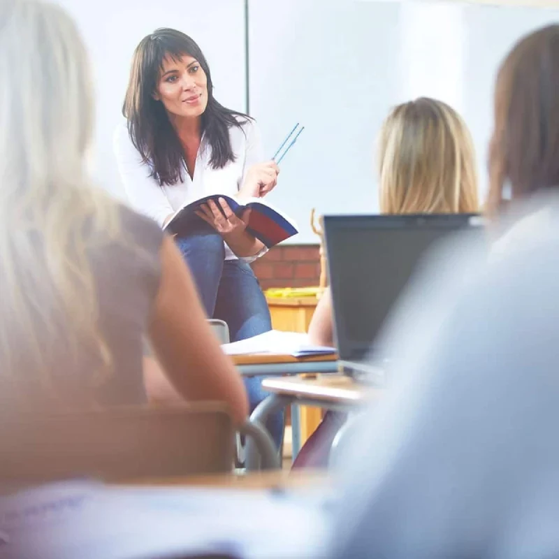 A teacher stands in front of a classroom, engaging students with a book in hand, while others listen attentively at their desks.