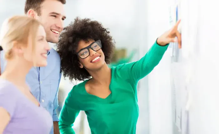 A woman in a green sweater gestures towards a wall with charts, while two colleagues observe her presentation attentively.