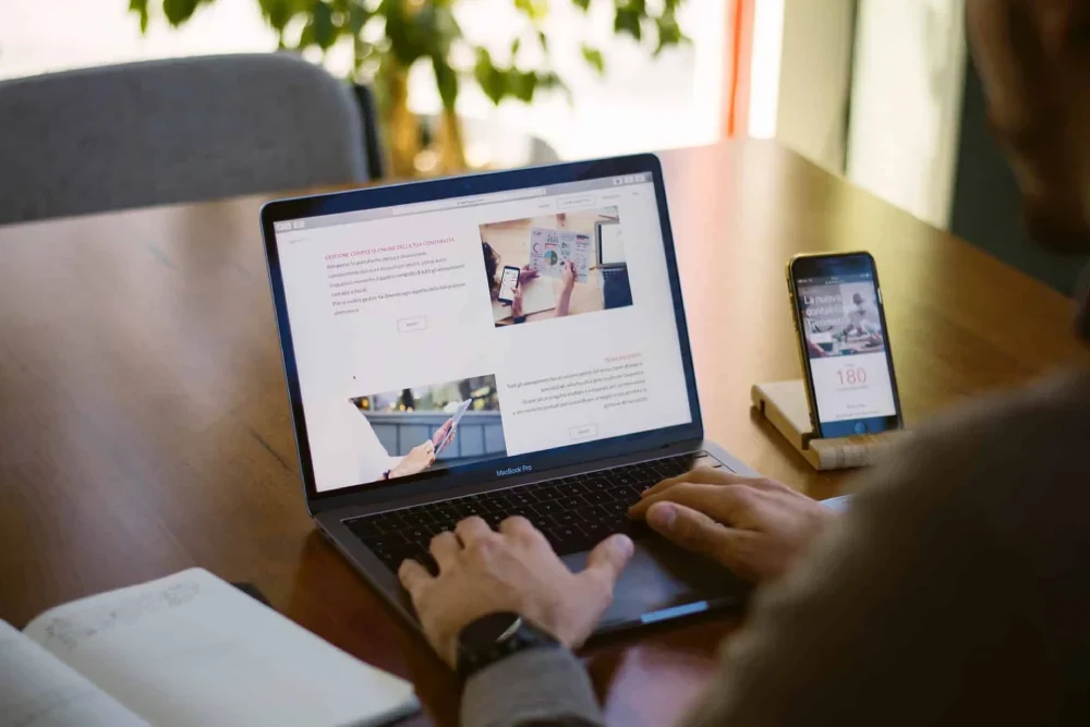 A person using a laptop to view a website, with a smartphone beside it on a wooden table in a well-lit room.